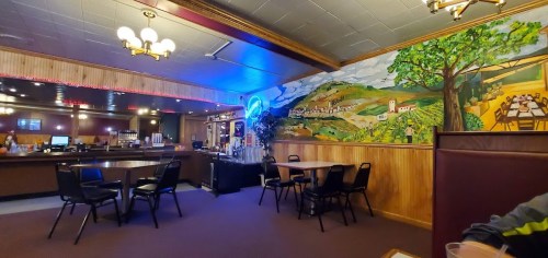 Interior of a restaurant featuring a mural of a vineyard, tables, chairs, and a bar area with soft lighting.