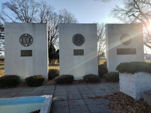 Three stone monuments with emblems and plaques, surrounded by shrubs, in a park setting with trees in the background.