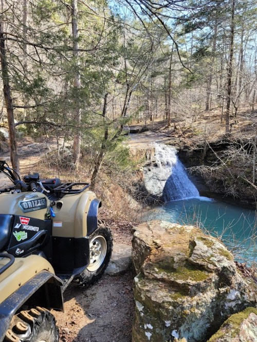 An ATV parked near a rocky edge overlooking a serene waterfall and turquoise water, surrounded by trees.