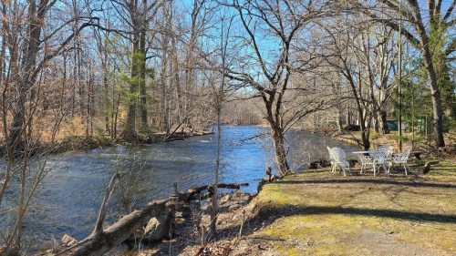 A serene river scene with bare trees, a grassy bank, and a small seating area by the water under a clear blue sky.
