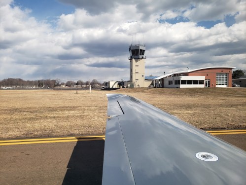 View from an airplane wing, showing a control tower and airport buildings under a cloudy sky.