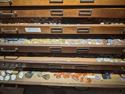 A wooden cabinet with multiple drawers filled with various colorful stones and minerals, labeled for identification.