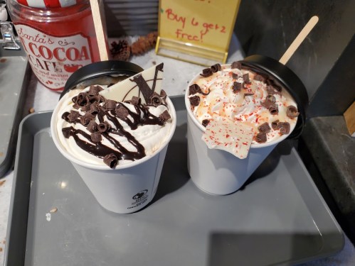 Two cups of festive hot chocolate topped with whipped cream, chocolate shavings, and crushed peppermint, on a tray.