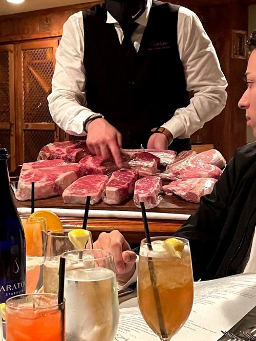 A waiter in a black vest points at a selection of raw meats on a table, with drinks and a menu in the foreground.