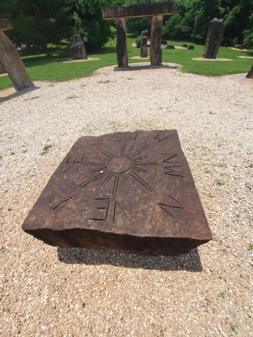 A large, engraved stone with a compass design sits on gravel, surrounded by tall standing stones in a green park.