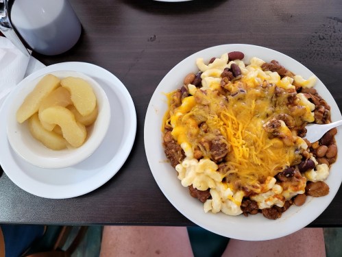 A plate of macaroni topped with chili, beans, and cheese, alongside a bowl of sliced peaches.