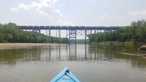 A kayak view of a river with a train bridge overhead, surrounded by lush greenery and a clear blue sky.