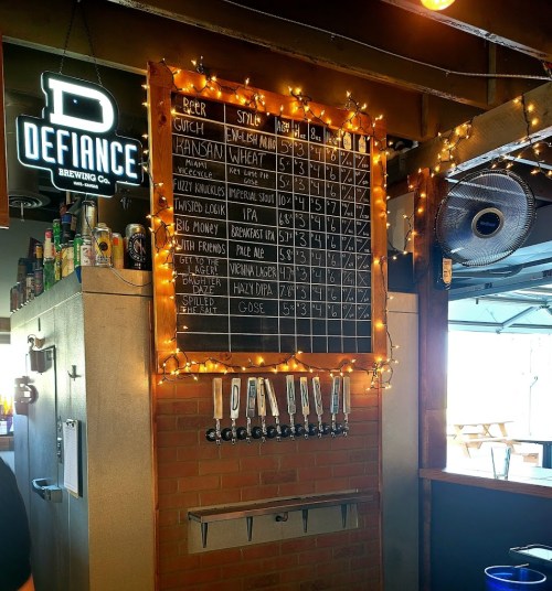 A chalkboard menu displaying various beers and styles, adorned with string lights in a brewery setting.