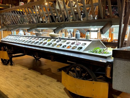 A long salad bar on a wooden cart, featuring various bowls of fresh vegetables and toppings in a rustic setting.