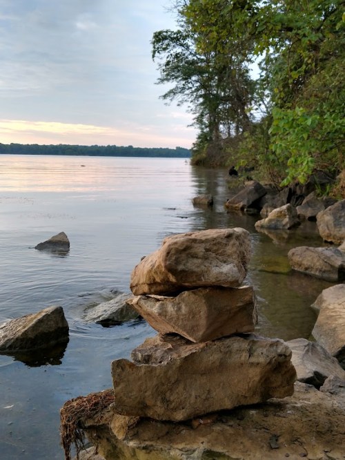 A stack of rocks by a calm river, surrounded by trees and reflecting the soft colors of the sky at sunset.