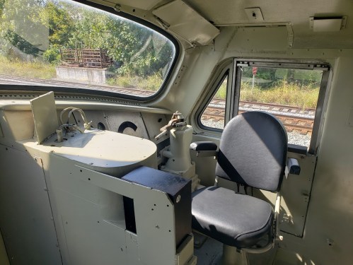Interior of a train cab featuring a control panel and a black seat, with a view of greenery and train tracks outside.