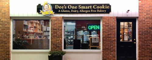 Exterior of Dee's One Smart Cookie bakery, featuring a sign and an "OPEN" neon light in the window.