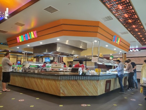 A bustling food court with a Mongolian grill and sushi station, featuring colorful signage and customers in line.