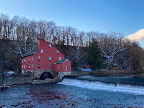 A red historic mill beside a river, with a waterfall and bare trees in the background under a blue sky.