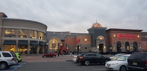 Exterior view of a shopping center featuring Cheesecake Factory and other stores, with cloudy skies above.