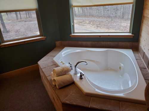 A cozy bathroom featuring a large, white jacuzzi tub with towels neatly stacked nearby and windows overlooking a wooded area.