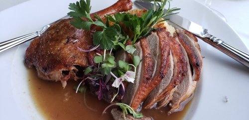 A plated dish featuring sliced duck breast, a whole duck leg, garnished with herbs and flowers, served with sauce.
