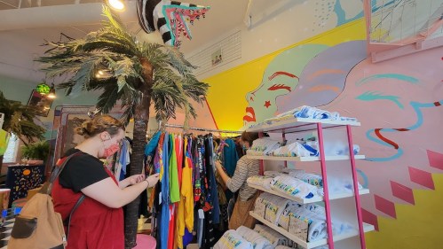 A colorful boutique with a palm tree, vibrant clothing racks, and a mural of a face on the wall. Shoppers browse the items.