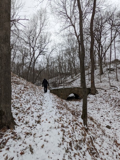 A person walks up a snowy path beside a stone bridge in a wooded area with bare trees.