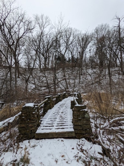 A snow-covered stone bridge leads through a winter landscape with bare trees and a hillside in the background.