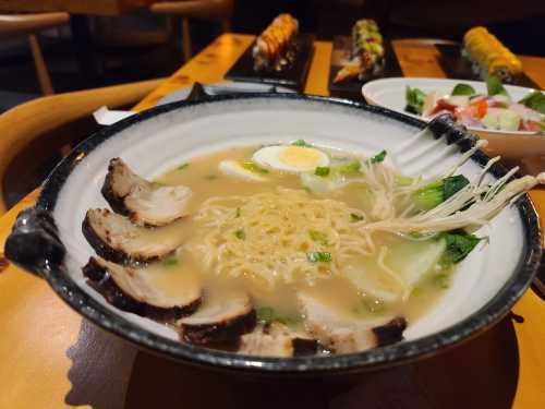 A bowl of ramen with sliced pork, egg, and green onions, served with sushi rolls and a side salad in the background.