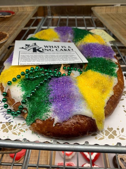 A colorful King Cake with green, purple, and yellow sugar, adorned with beads and an informational card.