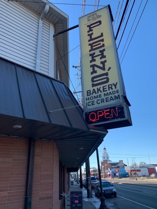 Sign for a bakery and ice cream shop, with "OPEN" illuminated, located on a street with a clear blue sky.