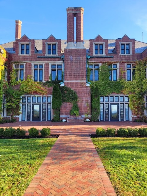 A brick mansion with ivy-covered walls, large windows, and a pathway leading to the entrance, set against a clear blue sky.