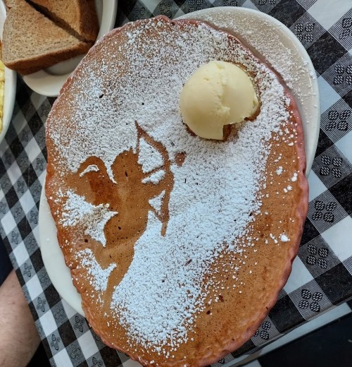 A large pancake dusted with powdered sugar, featuring a cupid design, topped with a scoop of ice cream.