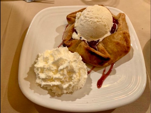 A dessert plate featuring a warm fruit pastry topped with vanilla ice cream and a side of whipped cream.