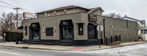 Exterior view of a modern restaurant building with large windows and a sign reading "Roux" on a cloudy day.