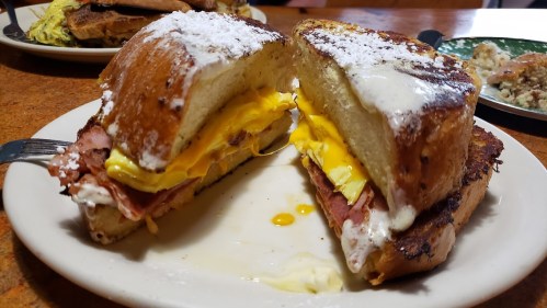 A close-up of a sandwich with layers of egg, ham, and creamy filling, served on toasted bread with powdered sugar.