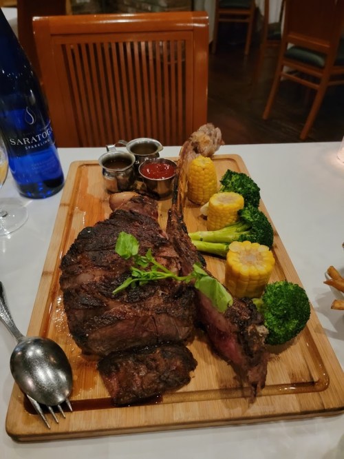 A beautifully plated steak with sides of corn, broccoli, and sauces on a wooden board, with a bottle of water in the background.