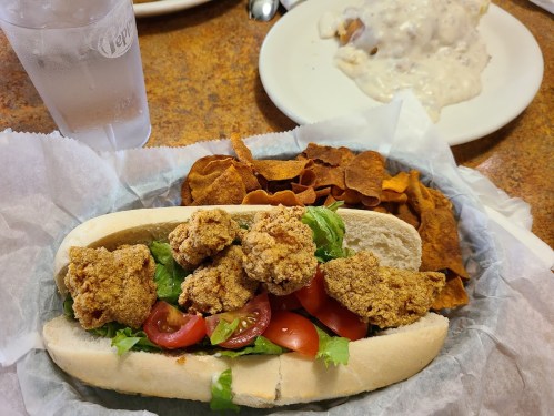 A sandwich with fried chicken, lettuce, and tomatoes, served with a side of crispy sweet potato chips and a glass of water.