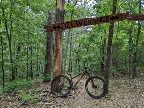 A mountain bike rests on a dirt path beneath a wooden sign reading "DELIVERANCE" in a lush green forest.