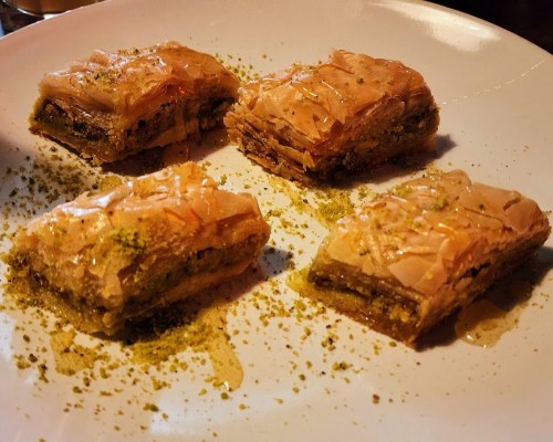 A plate of four pieces of baklava drizzled with honey and sprinkled with crushed pistachios.