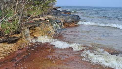 A rocky shoreline with gentle waves lapping against the shore, surrounded by greenery and a calm blue sea.