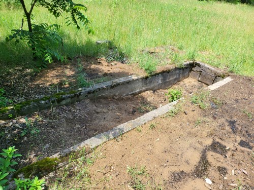 A partially overgrown concrete foundation in a grassy area, surrounded by sparse vegetation and dirt.