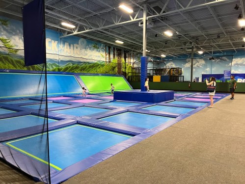 A spacious indoor trampoline park with multiple trampolines and a central landing area, featuring colorful mats and walls.
