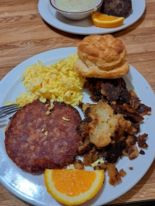A plate of breakfast featuring scrambled eggs, hash browns, a biscuit, a sausage patty, and an orange slice.