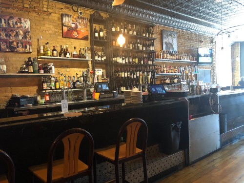 A cozy bar with a wooden counter, shelves of liquor, and empty chairs in a warm, inviting atmosphere.