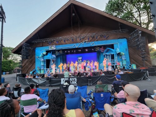 A colorful outdoor stage with performers in costumes, while an audience sits in folding chairs in front.