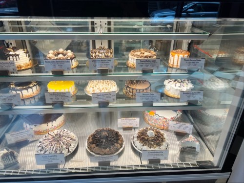 A display case filled with various decorated cakes, each labeled with names and descriptions.