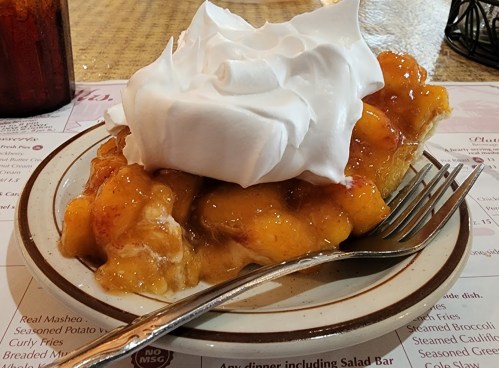 A slice of peach cobbler topped with whipped cream on a plate, with a fork beside it.
