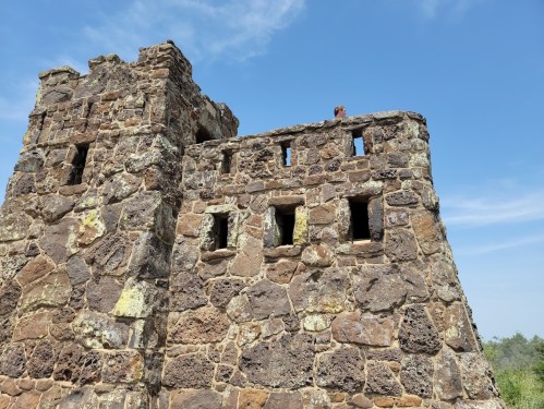 A weathered stone tower with small windows against a clear blue sky, surrounded by greenery.