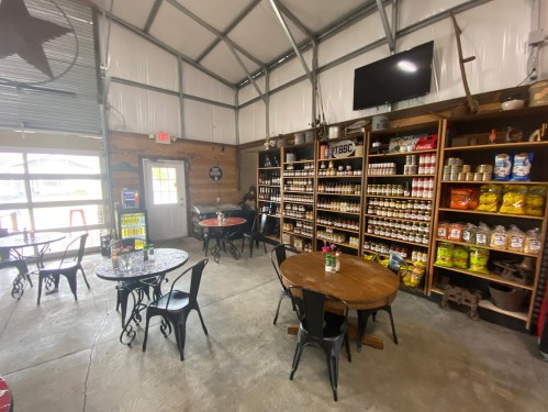 Cozy interior of a rustic shop with tables, shelves of goods, and a TV, featuring a welcoming atmosphere.