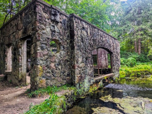 A weathered stone structure beside a serene stream, surrounded by lush greenery and trees.