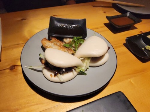 A plate of bao buns filled with grilled meat and garnished with herbs, served with dipping sauces on a wooden table.