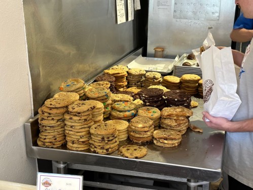 A table piled high with various stacks of cookies, with a person holding a bag nearby.