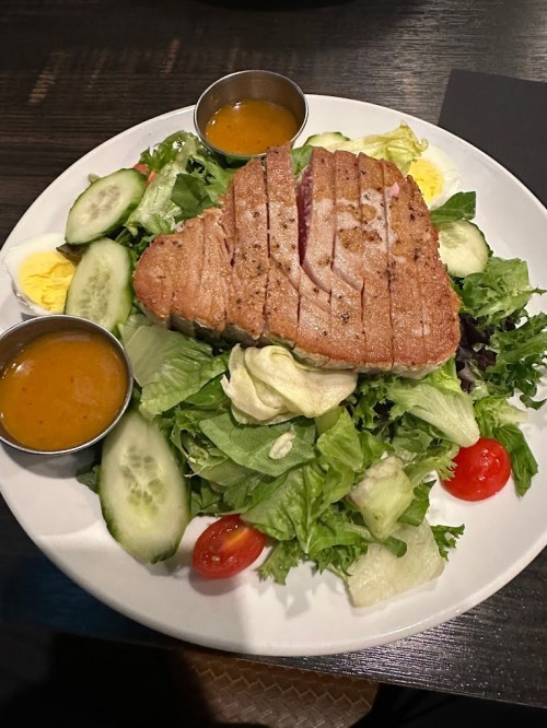 A plate of salad topped with grilled tuna, sliced cucumbers, cherry tomatoes, and two small cups of dressing.
